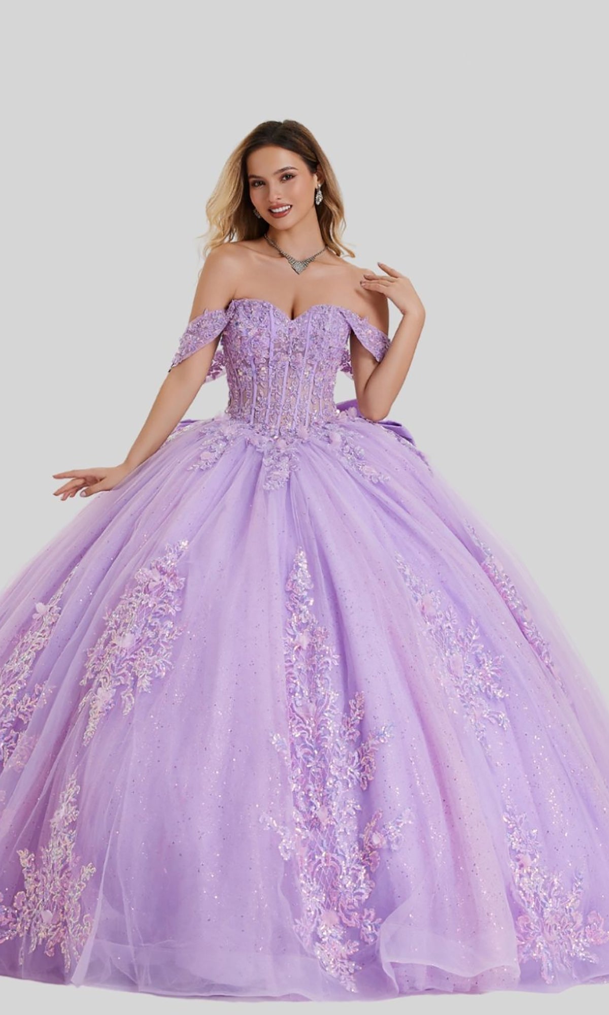 A woman wearing the Quinceanera Dress 26408 by Stella Couture in lilac, featuring intricate floral embroidery, shimmering sequins, and a full tulle ball gown skirt, stands against a plain light gray background.
